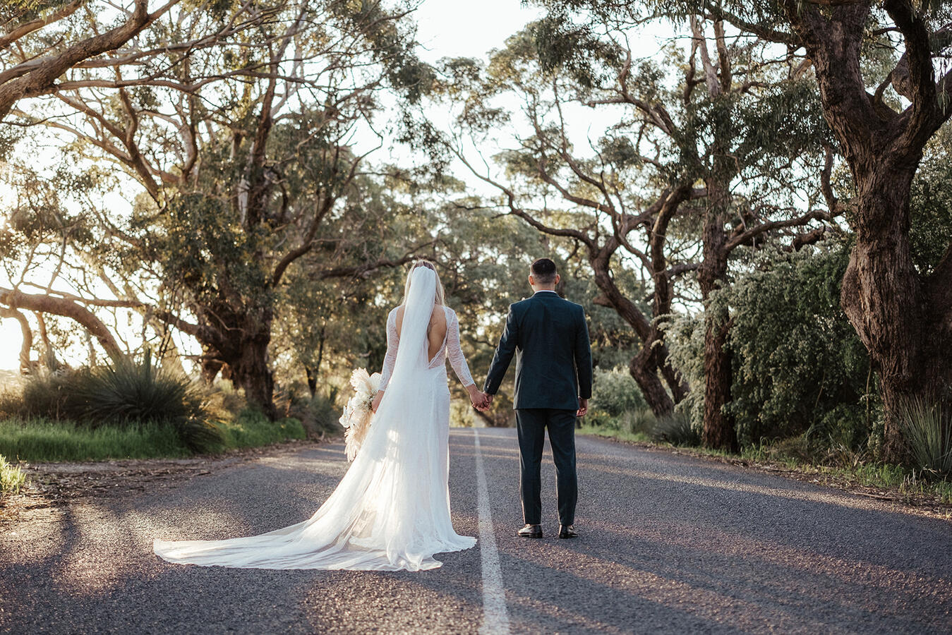 A bride and groom holding hands on a country road surrounded by trees.
